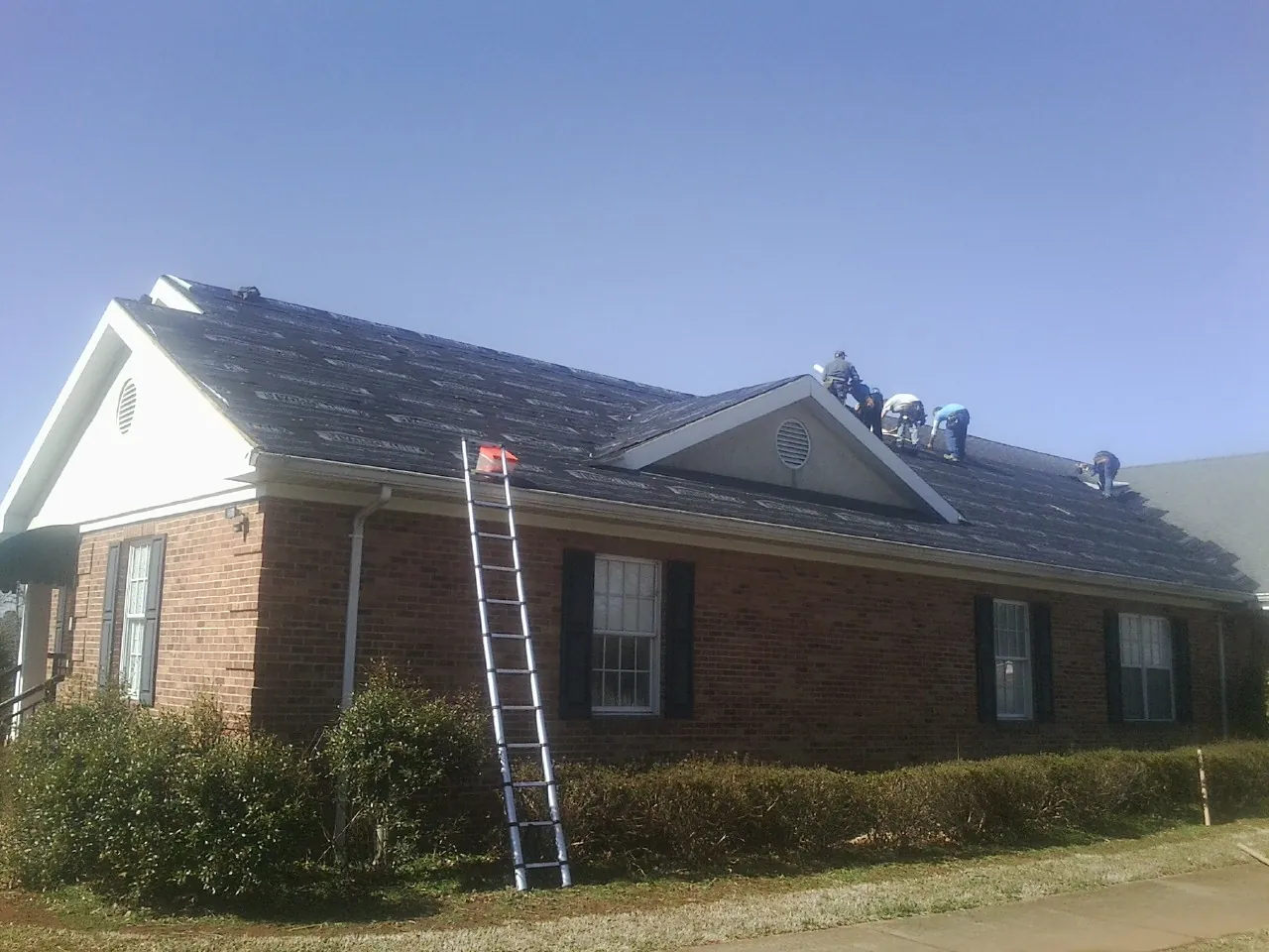 Crew tearing shingles off a brick ranch roof with a ladder set — Metal Roof Repair work by our crew in East Marion