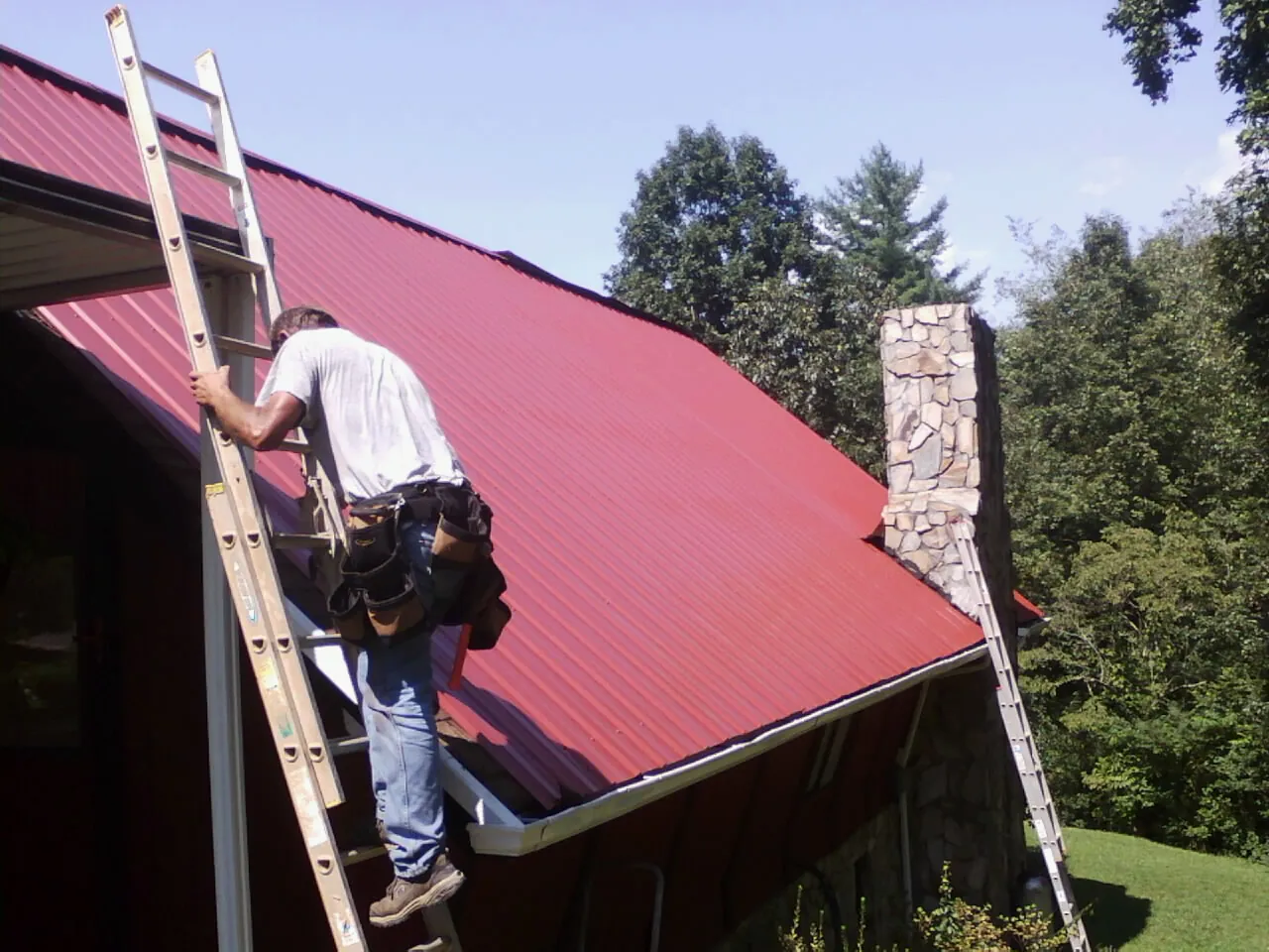 Roofer on a ladder beside a red metal roof and stone chimney — Soffit & Fascia Repair by Roofing Brothers in East Marion